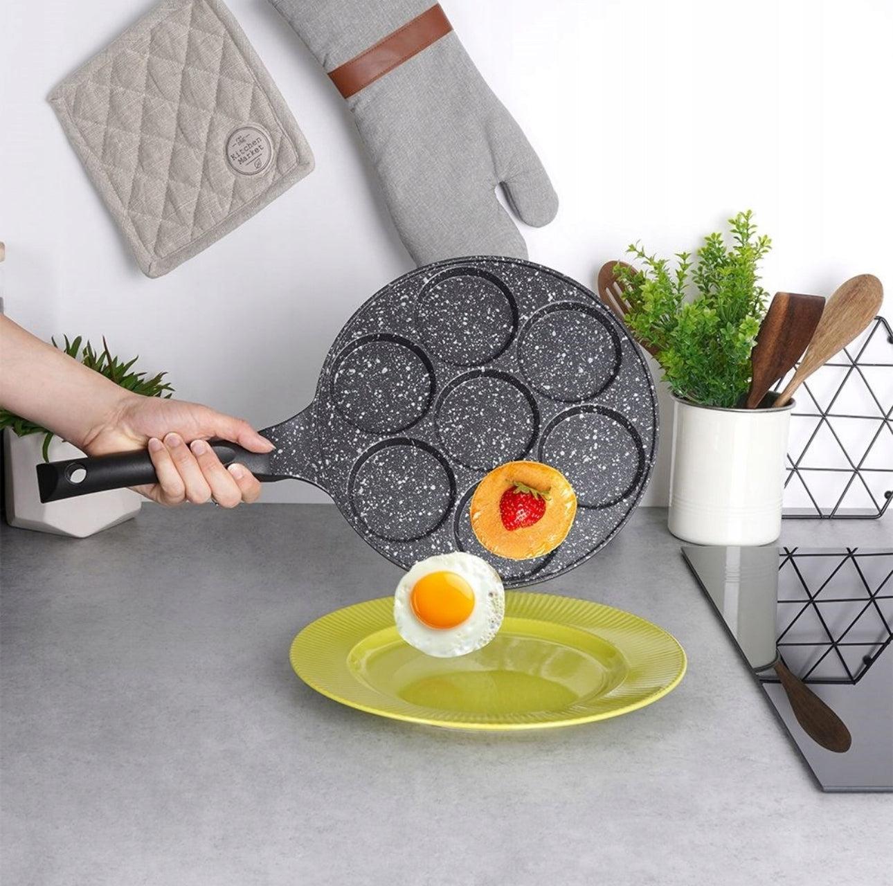 Hånd som heller egg og pannekake fra stekepanne i svart granittlook mot gul tallerken