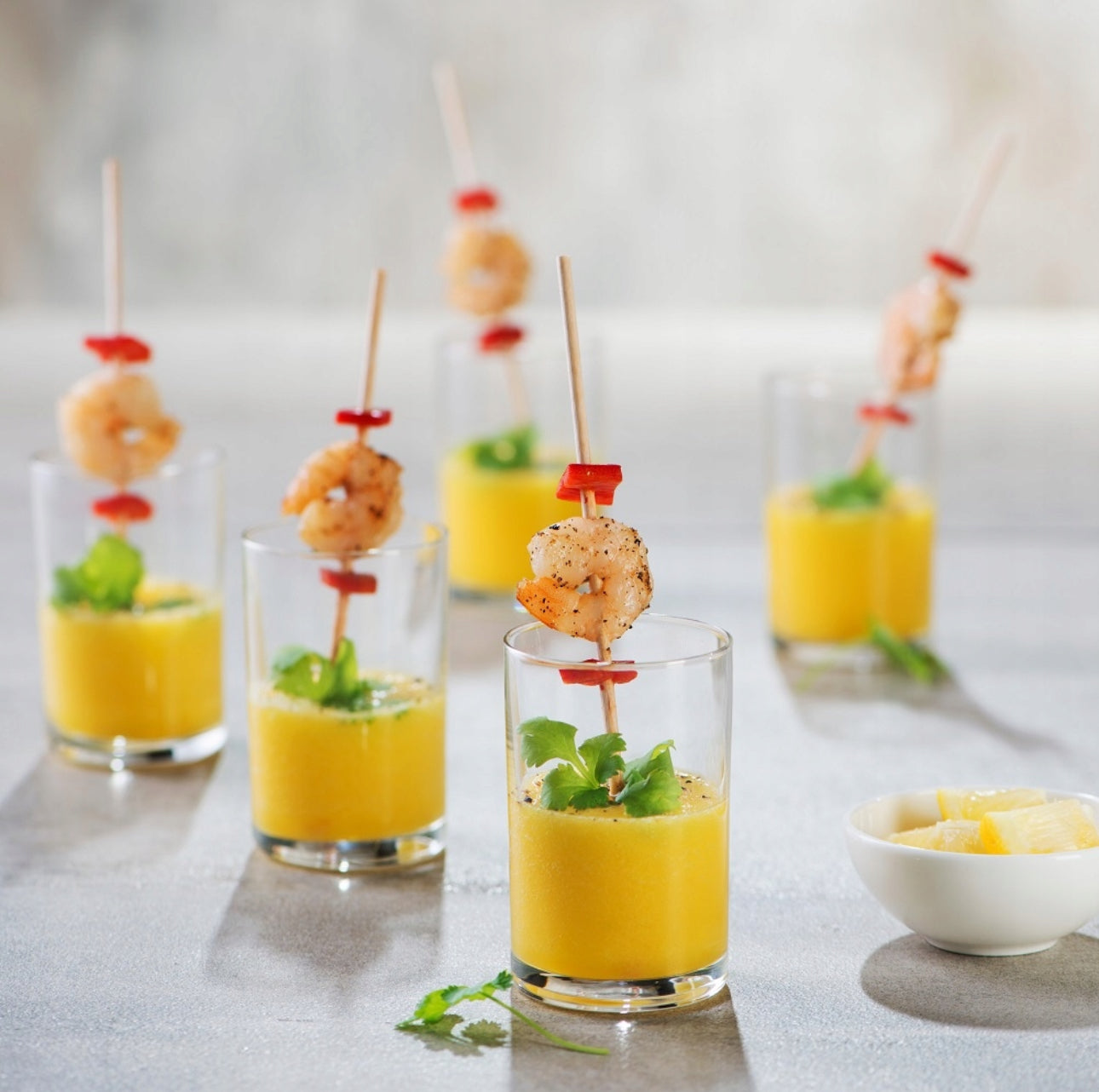 Små drikkeglass fylt med gul smoothie servert med reker og urter på spyd