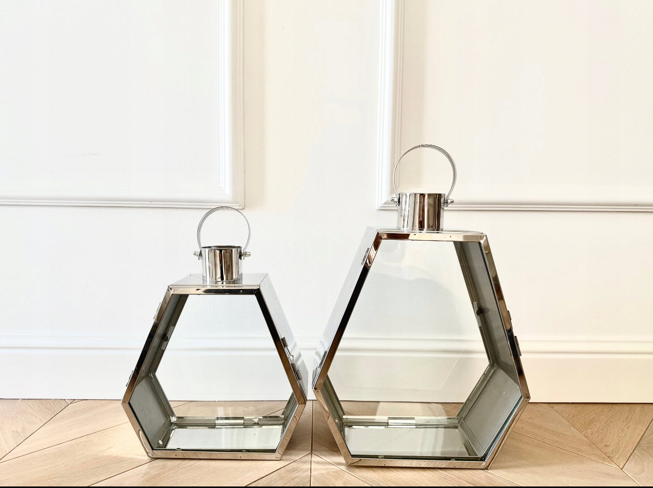 Sølv lykter i stål og glass stående på tregulv mot hvit veggpanelering