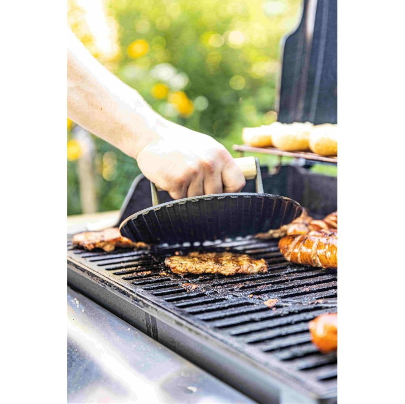Hånd holder grillpresse i støpejern over burger på utendørs grill med grønne omgivelser