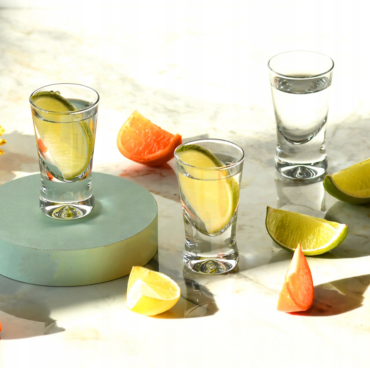 Tre shotglass fylt med vann og limebåter på marmorbord med frukt rundt