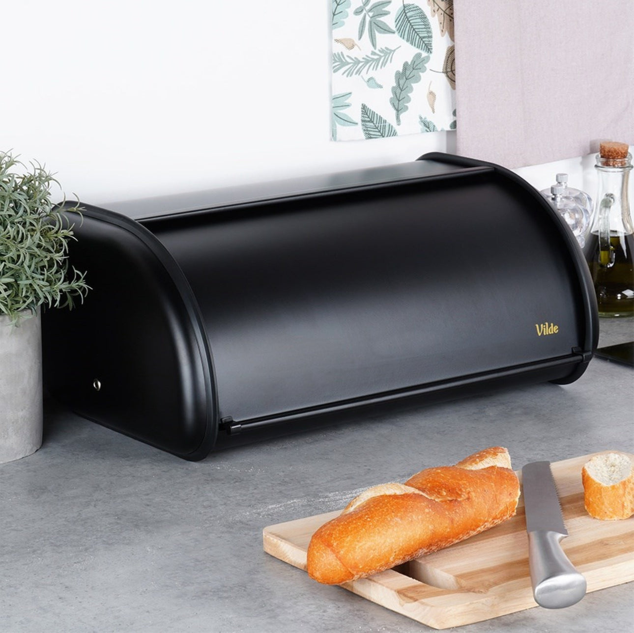 Svart brødboks på kjøkkenbenk omgitt av urter, skjærefjøl og baguette