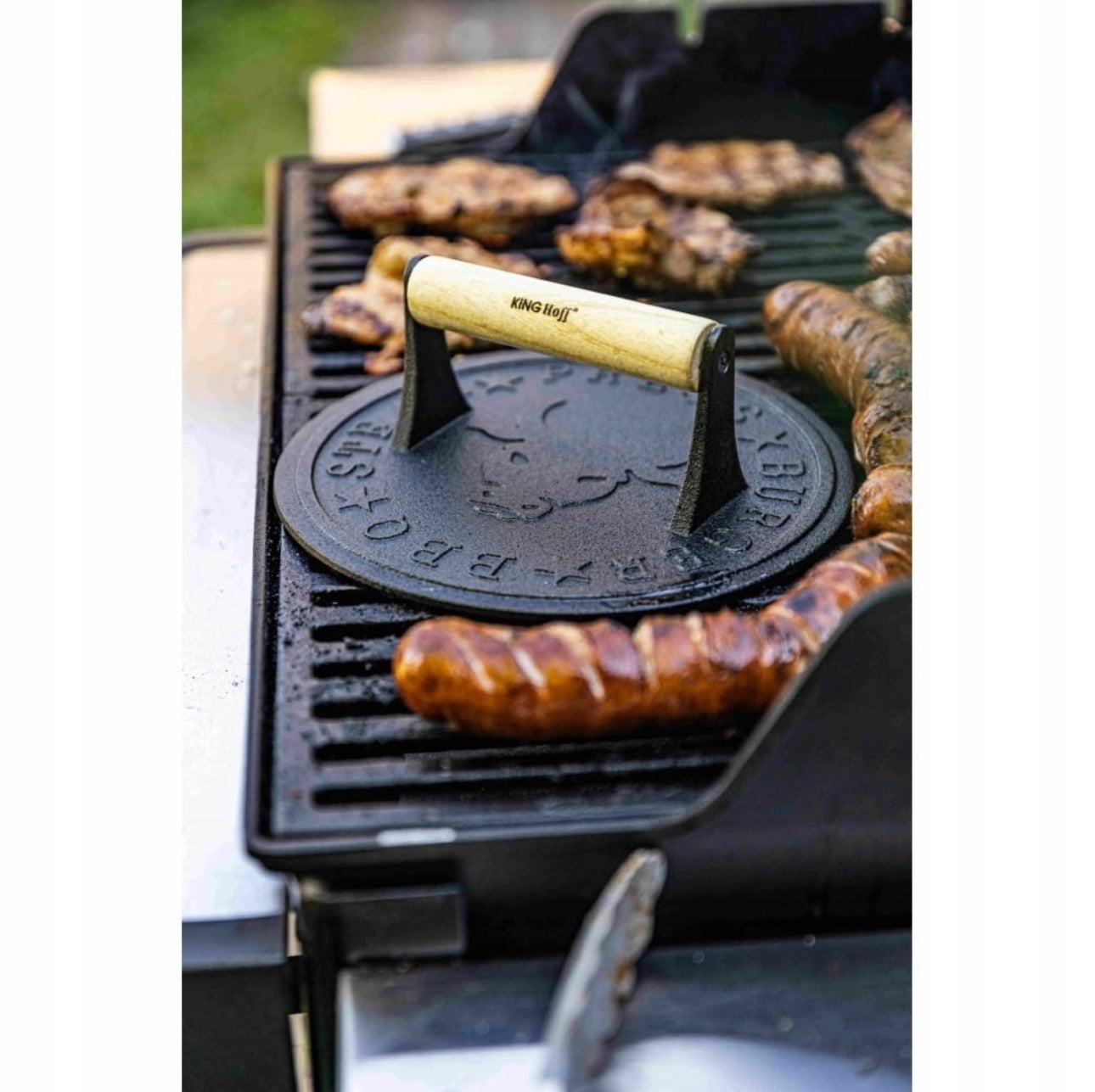 Burgerpresse med trehåndtak plassert midt på grillen blant pølser og kjøttstykker