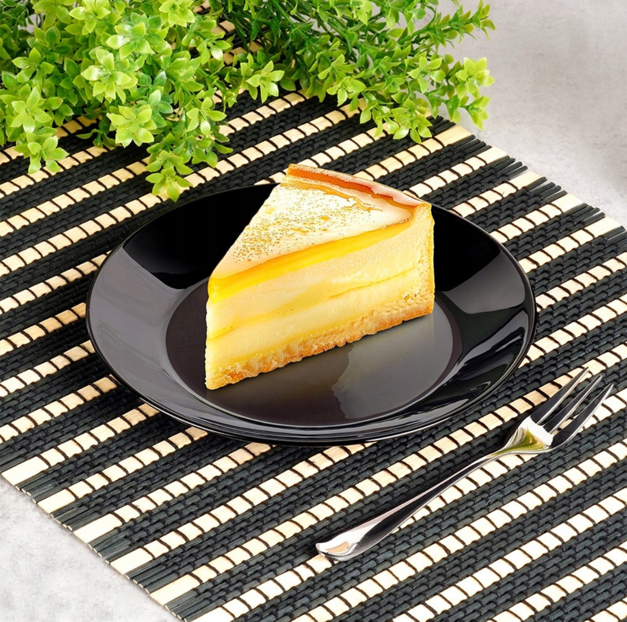 Svart desserttallerken med sitronkake, servert med gaffel på stråmatte med grønne planter