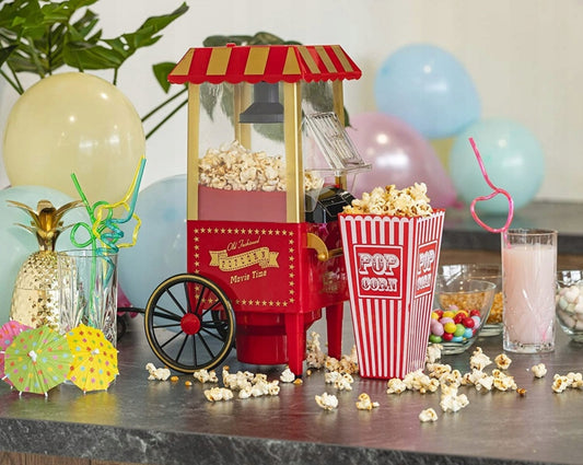 Rød retro popcornmaskin i plast med gull detaljer omgitt av popcorn, drikke og festdekor på kjøkkenbenk