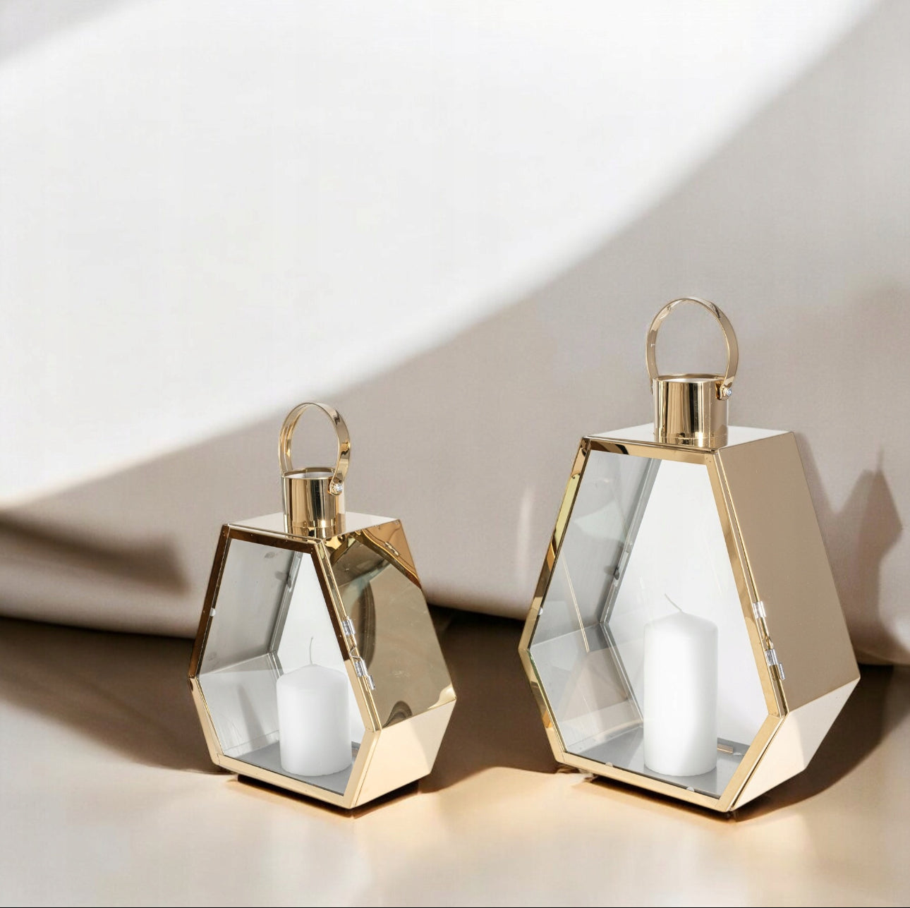 To moderne lykter i gull med hvite lys, stående på beige overflate i naturlig dagslys