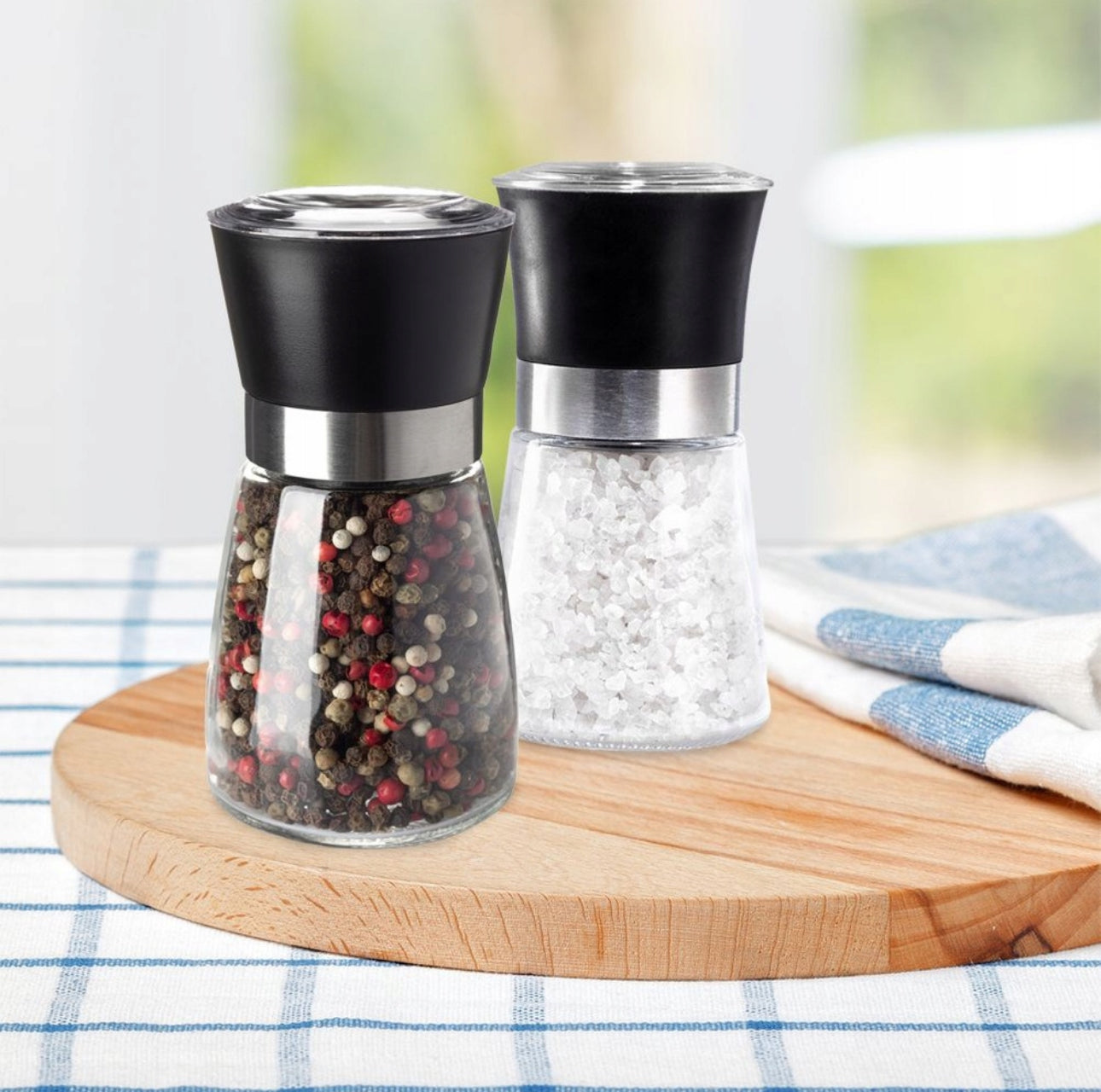 Salt- og pepperkverner i glass med sort topp, plassert på trebrett på kjøkkenbord med duk.