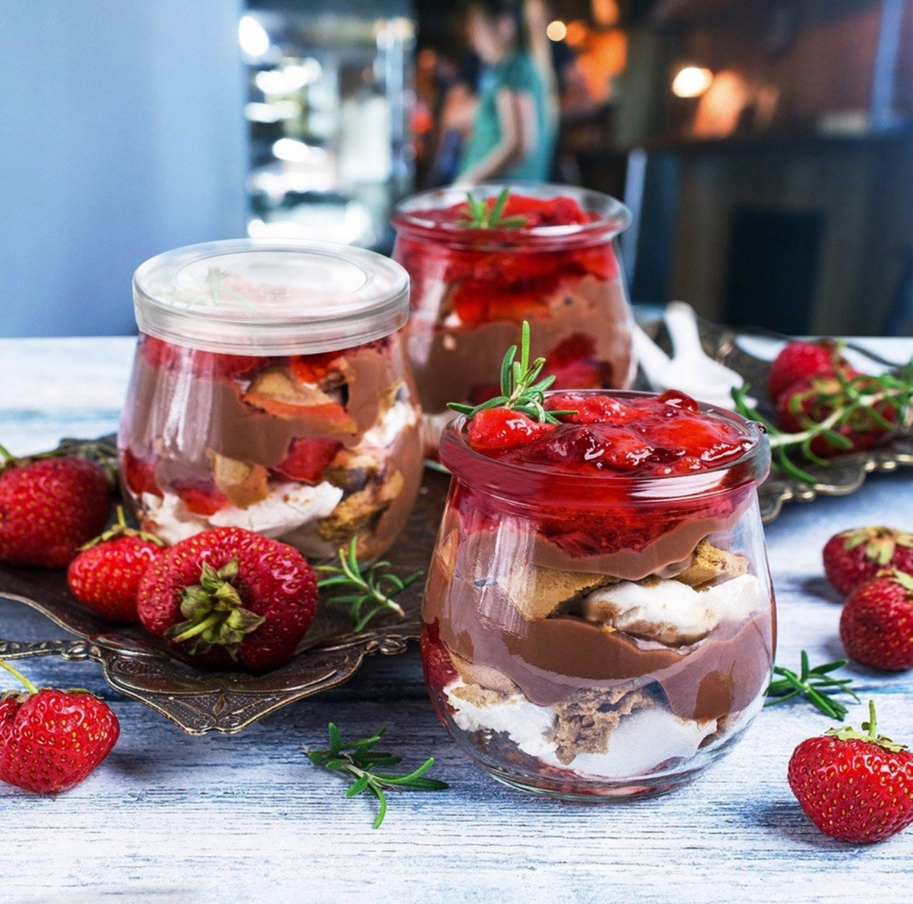 Dessertglass med sjokolade, krem og jordbær servert i glasskrukker på et bord med jordbær og urter