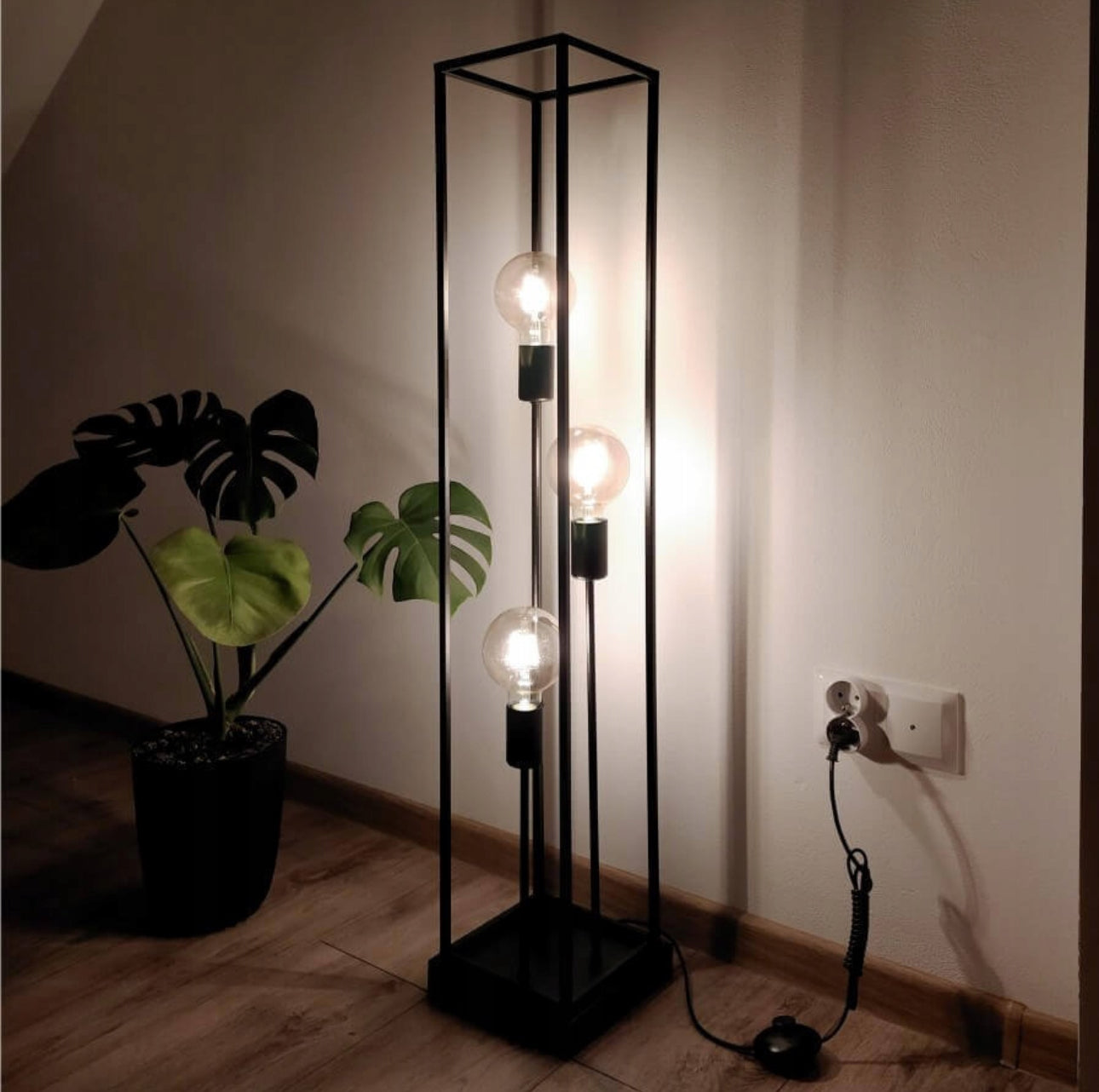 Tennes gulvlampe med tre lyspærer i svart metallramme ved grønn plante