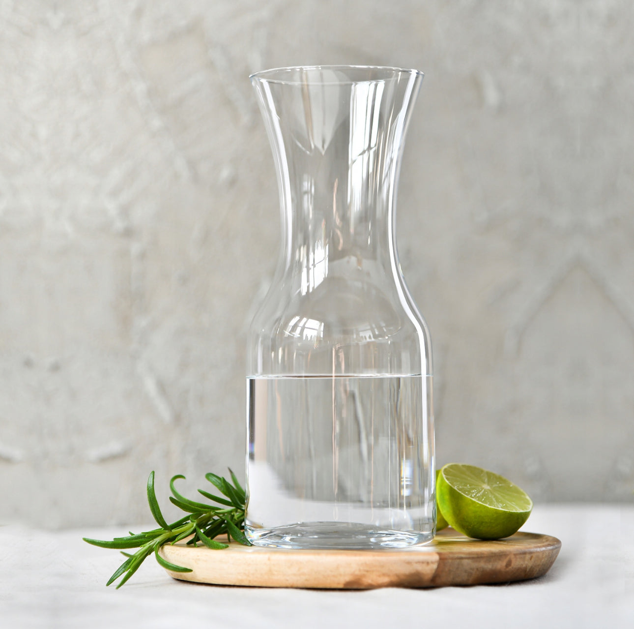 Elegant glasskaraffel med vann, lime og urter på trefat mot lys bakgrunn