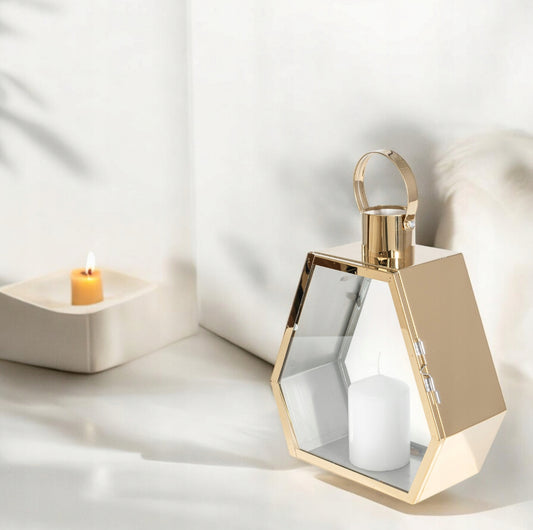 Gullfarget lykt i metall og glass med hvit kubbelys, plassert på hvit overflate ved vegg