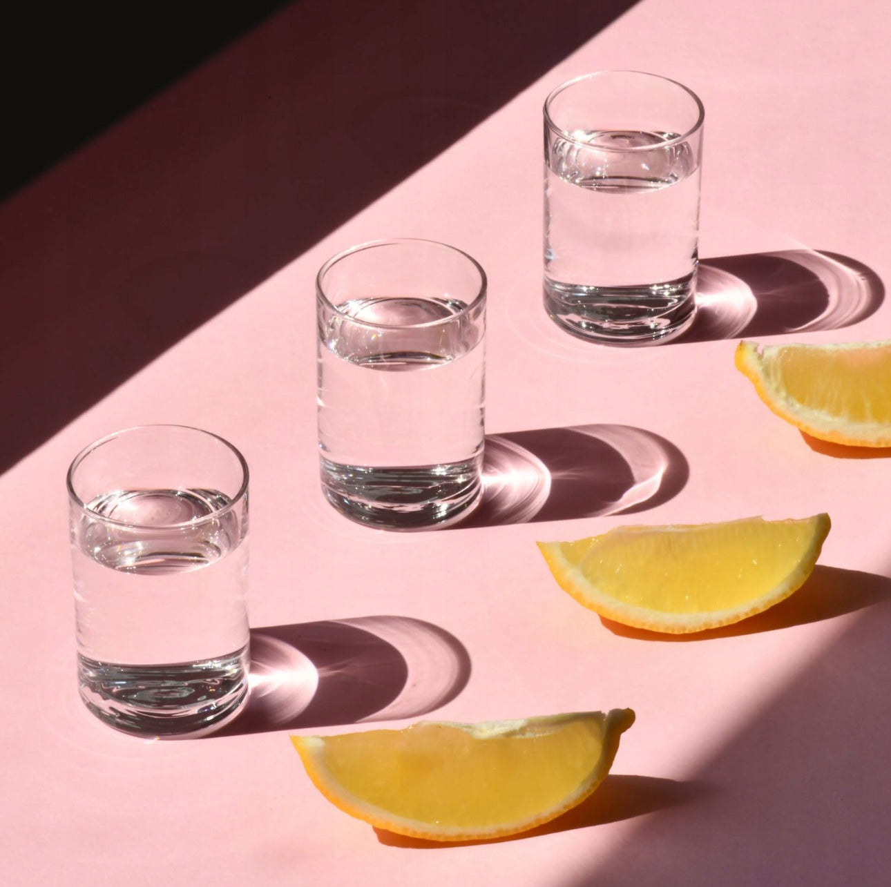 Tre shotglass med vann og sitronbåter på rosa bordflate