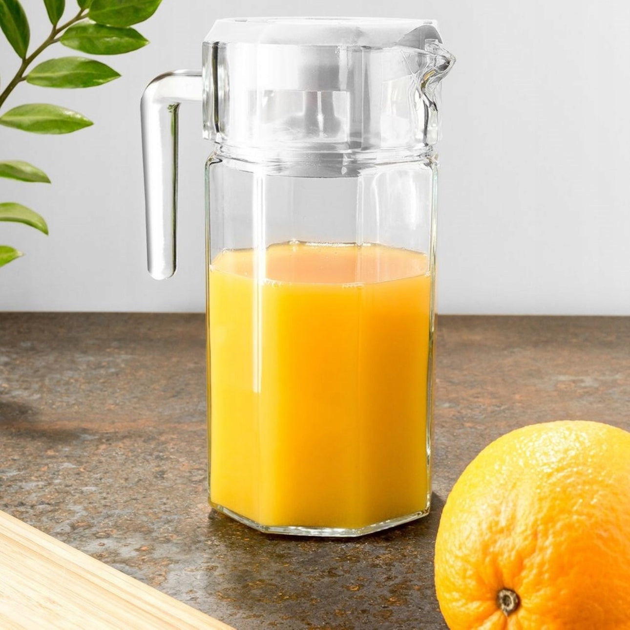 Serveringskanne med juice på kjøkkenbenk, omgitt av friske appelsiner og grønne blader