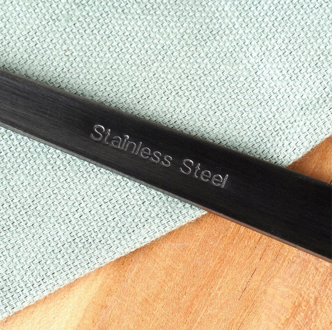 Gravert tekst “Stainless Steel” på mørkt salatbestikk mot tekstilbakgrunn