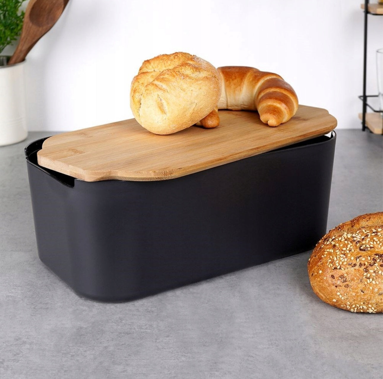 Svart brødboks med brød og croissant på bambuslokk i kjøkkenmiljø