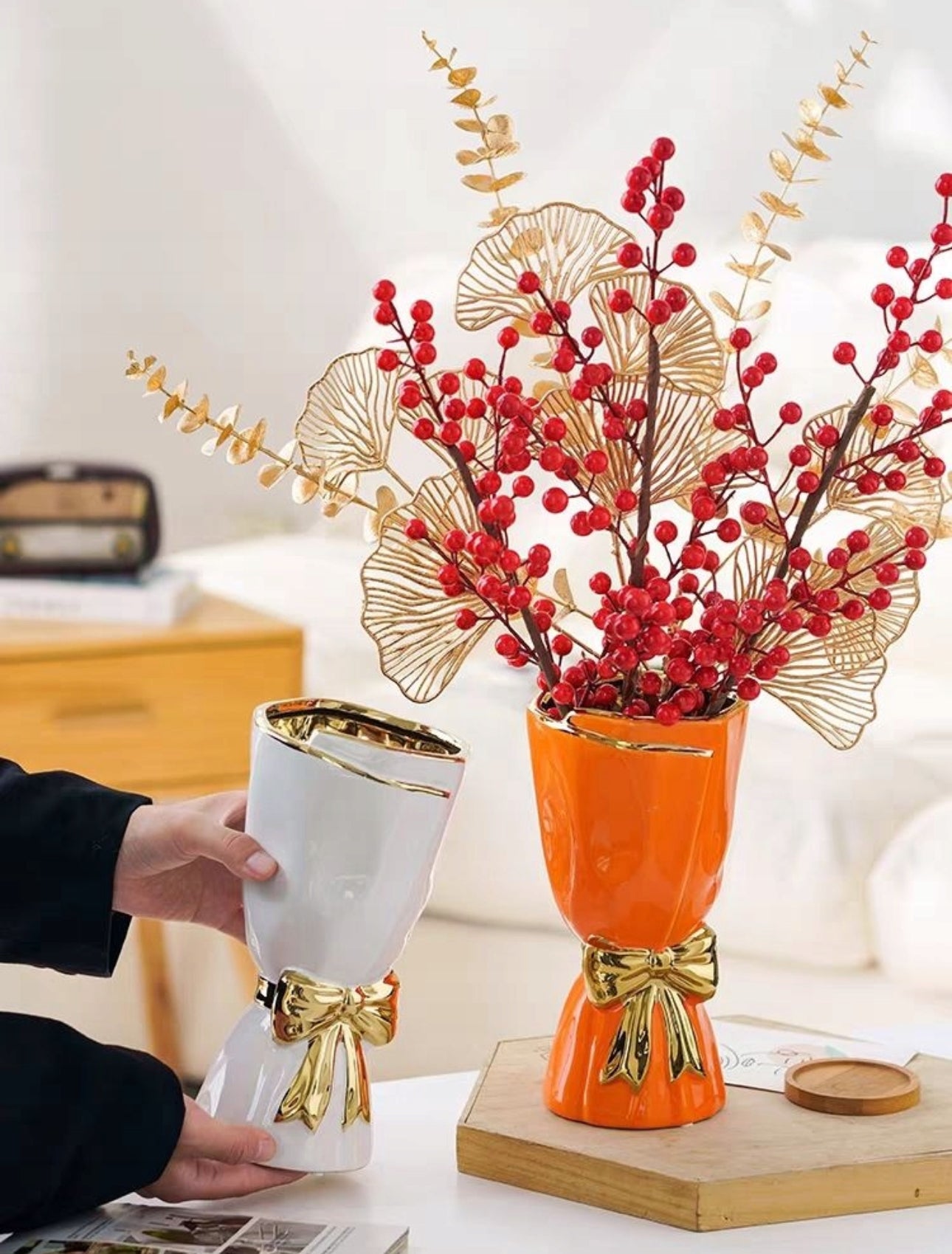 Oransje og hvit keramisk vase med gullsløyfe, plassert på bord med røde og gyldne dekorgrener