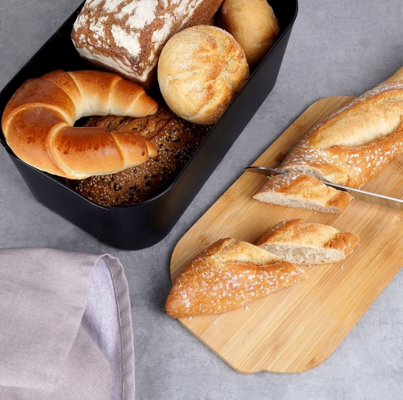 Skåret baguette på bambuslokk ved siden av svart brødboks med ferskt brød