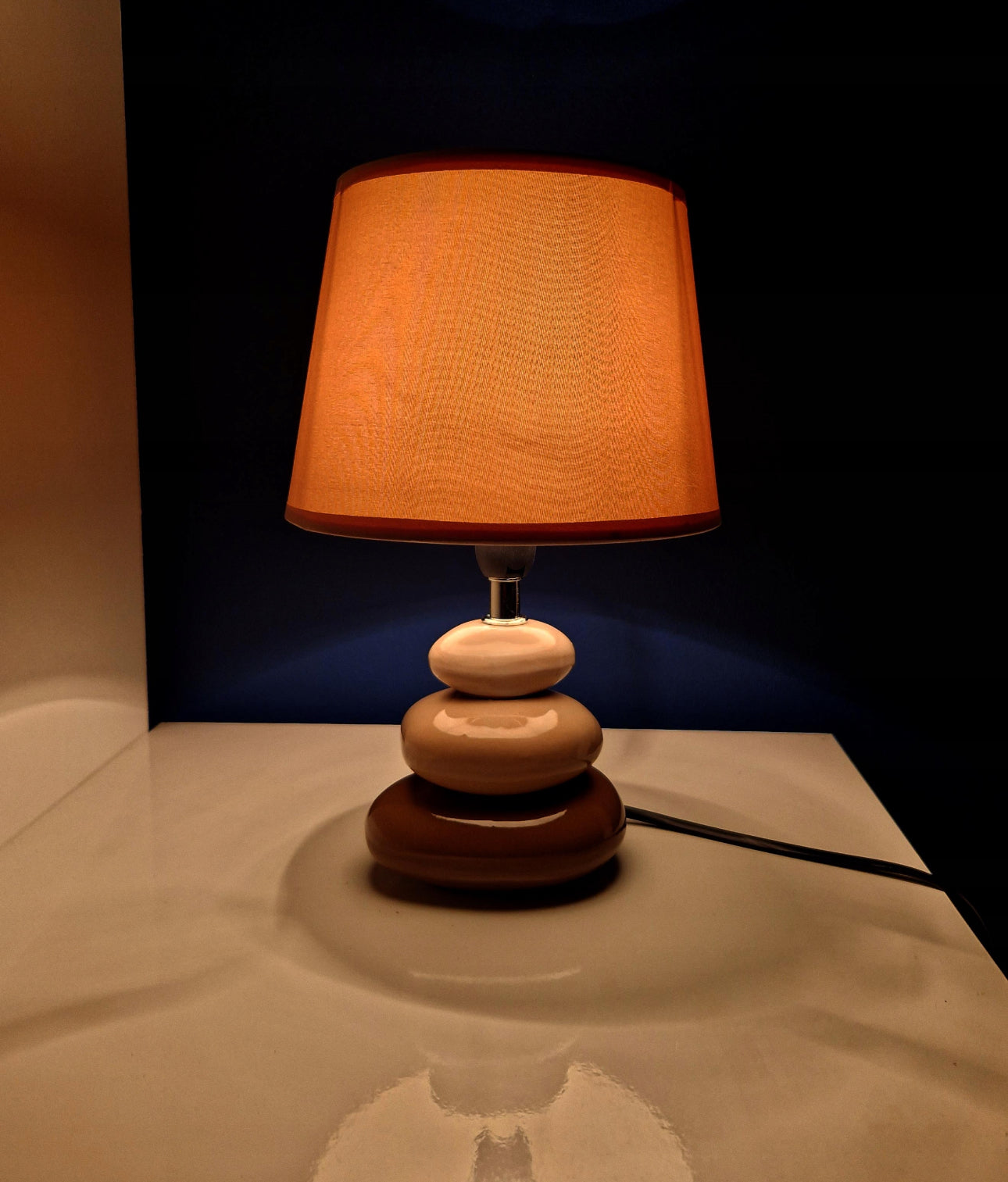 Tent nattbordslampe med beige skjerm som gir varmt lys i mørkt rom