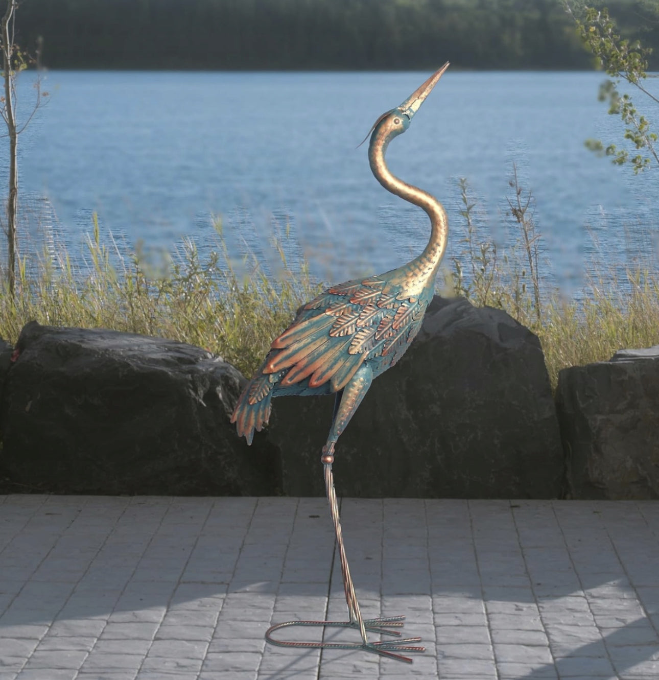 Kunstnerisk metalltrane i blå og kobbertoner, utstilt ved vannkanten med steiner i bakgrunnen.