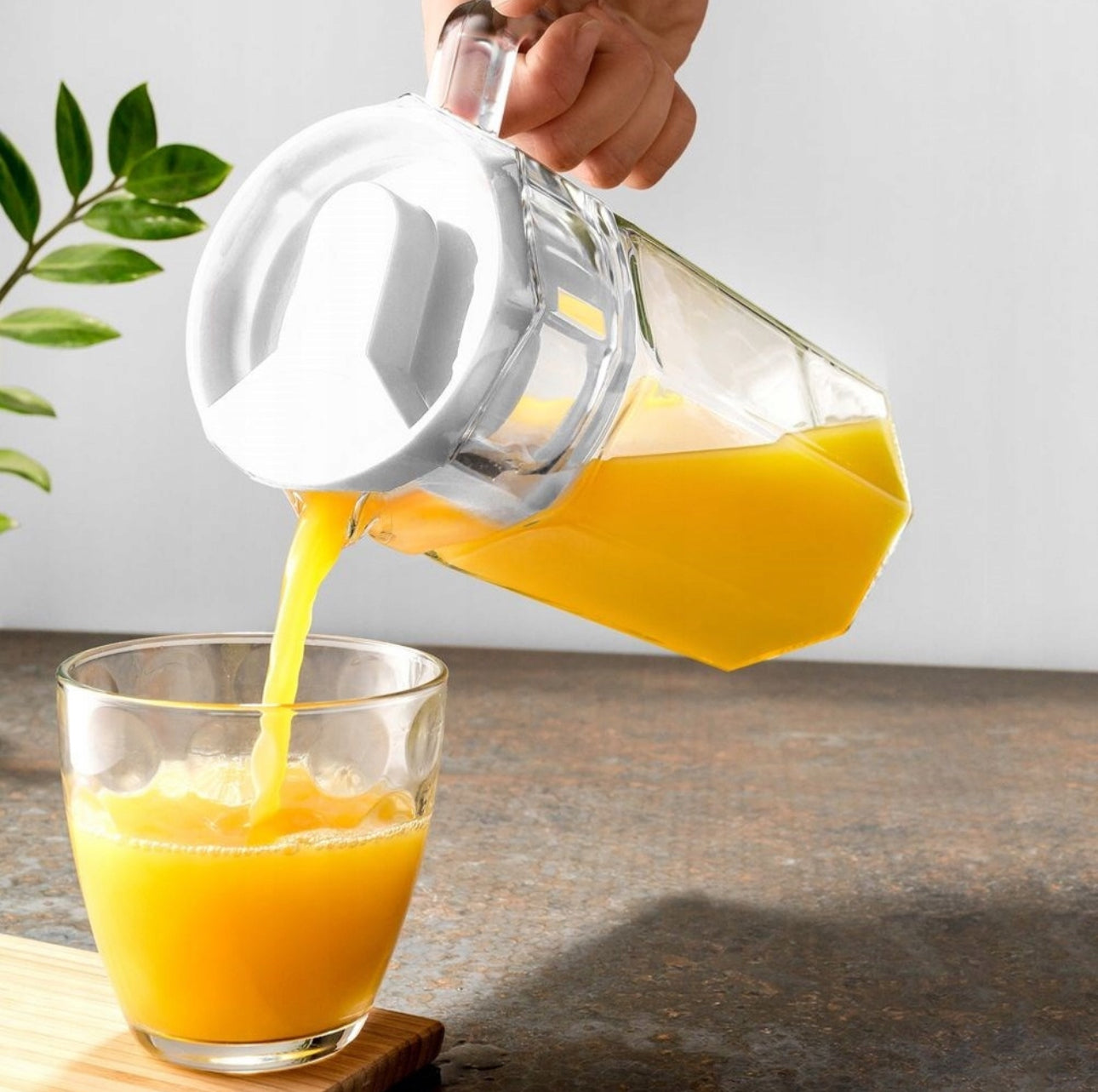 Juicekanne i bruk, helles i glass på benk med planter i bakgrunnen