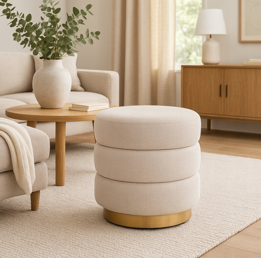 Elegant puff i kremfarget stoff med gyllen base, plassert i moderne stue