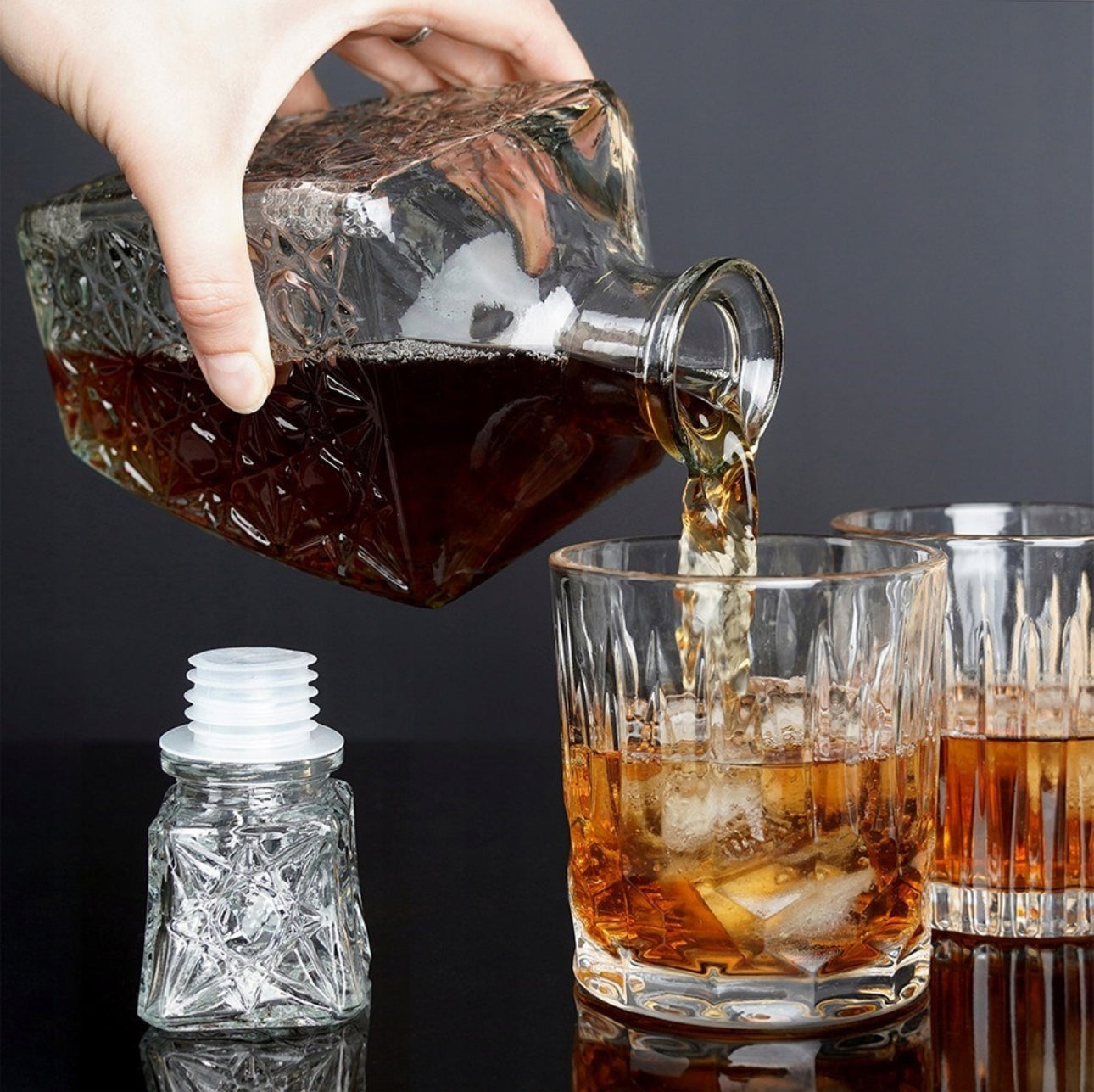 Hånd som heller brun væske fra mønstret glasskaraffel ned i whiskyglass med isbiter