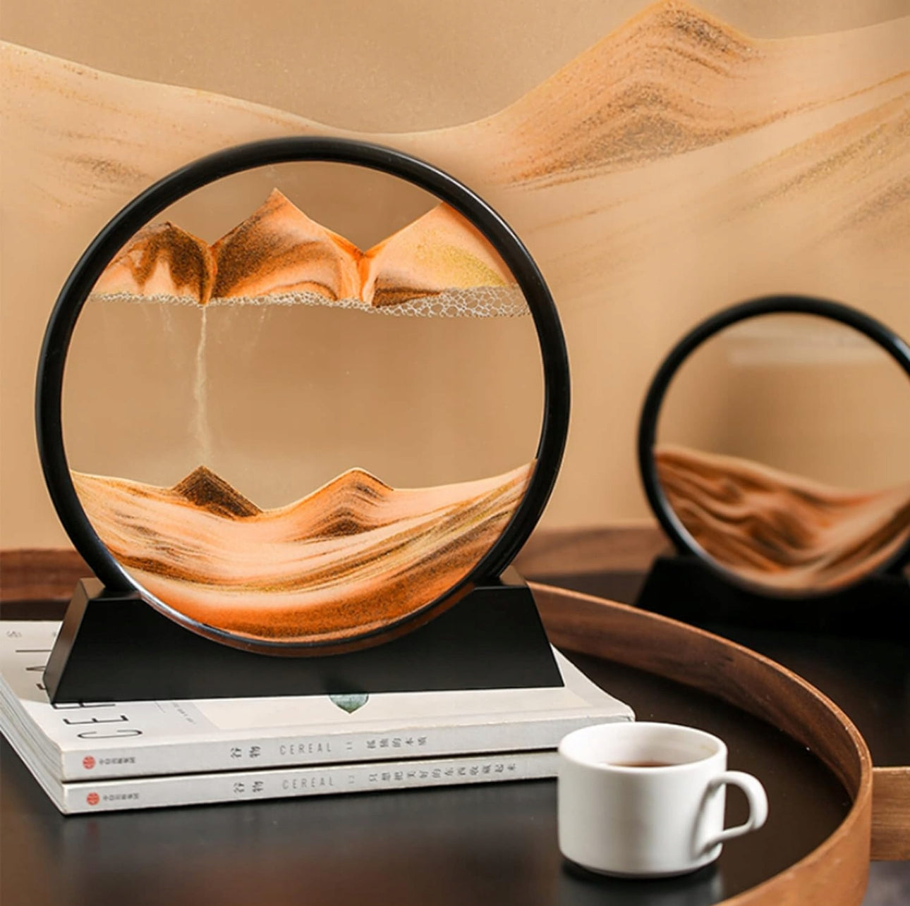 Oransje og brunt sandmaleri i rund ramme på svart base, plassert på bøker med kaffekopp ved siden av