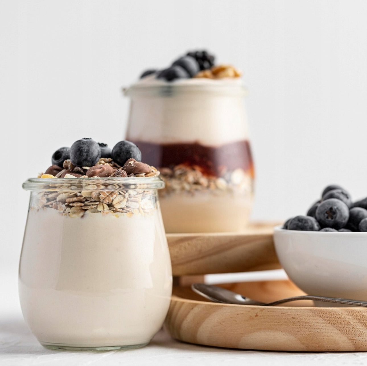 To glasskrukker med yoghurt, blåbær og müsli på lys bakgrunn med treserveringsbrett