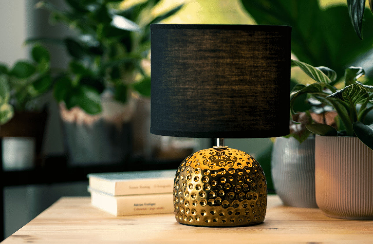 Glamour bordlampe med gullfarget base og svart skjerm plassert på trebord med grønne planter