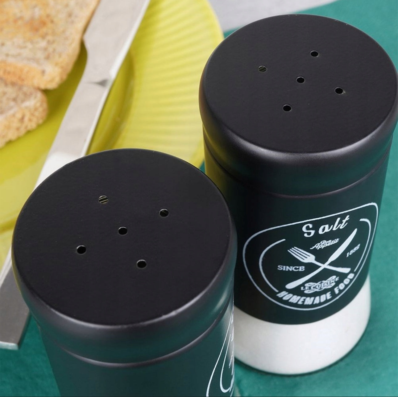 Nærbilde av toppen på salt- og pepperbøsser med ulikt antall hull i sort metall, plassert på frokostbord.