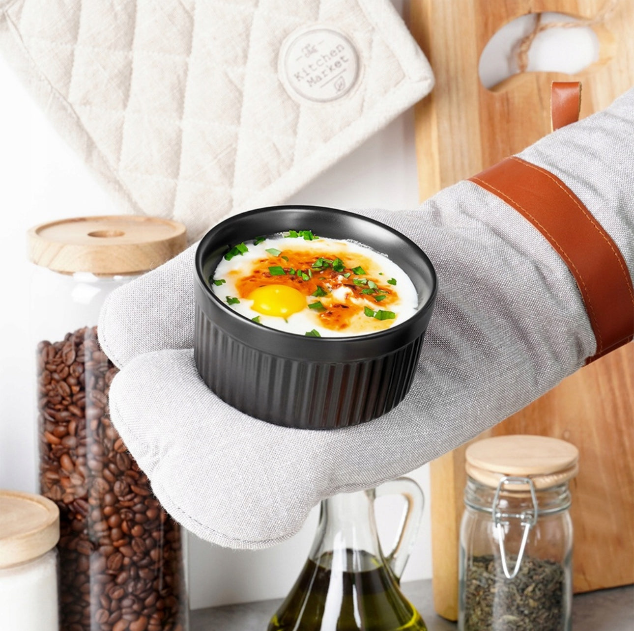 Svart keramikkform med egg og urter, holdt med grytevott i lys kjøkkeninnredning