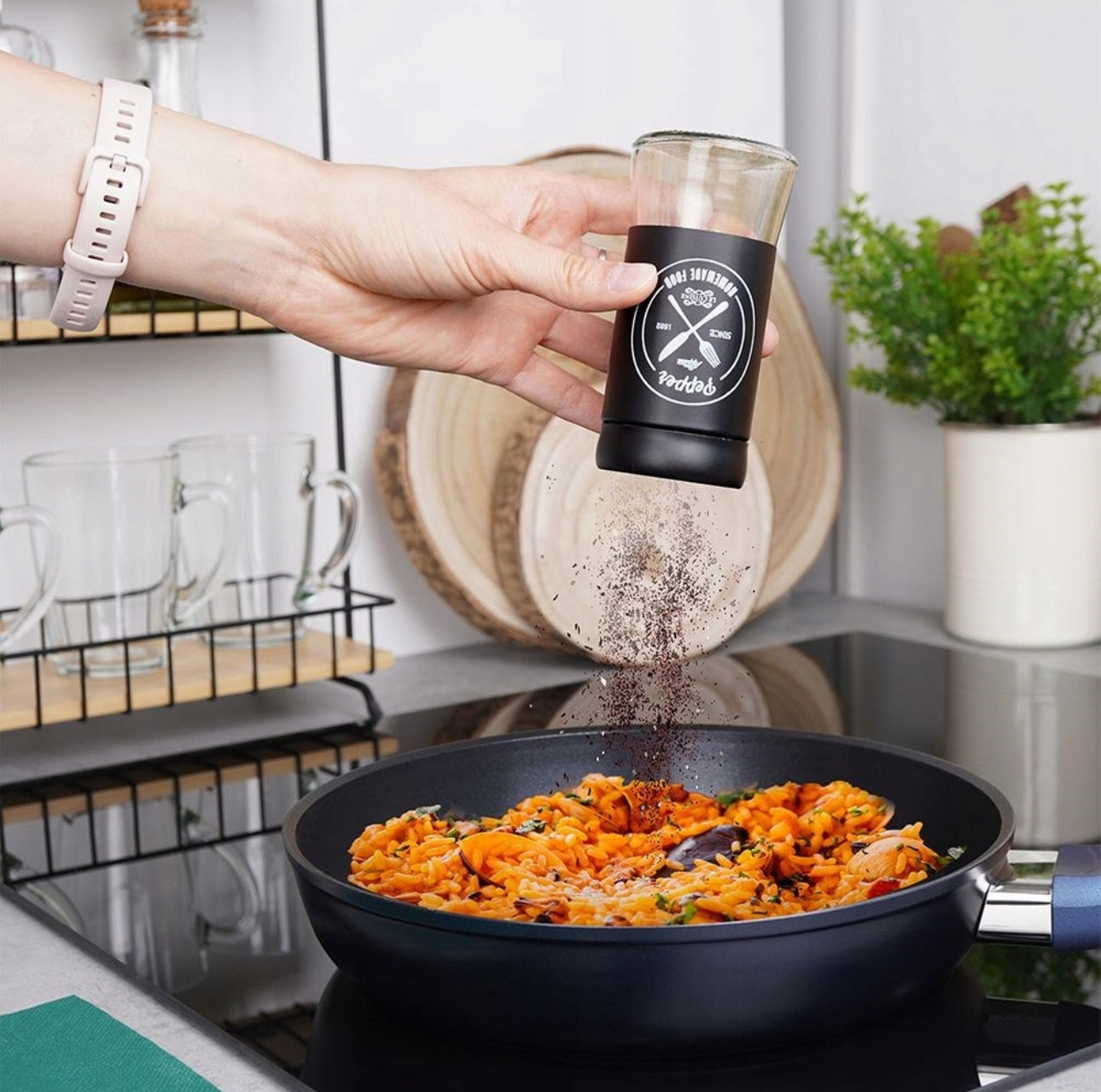 En hånd drysser pepper fra sort krydderbøsse over fargerik pastarett i stekepanne på moderne kjøkken.