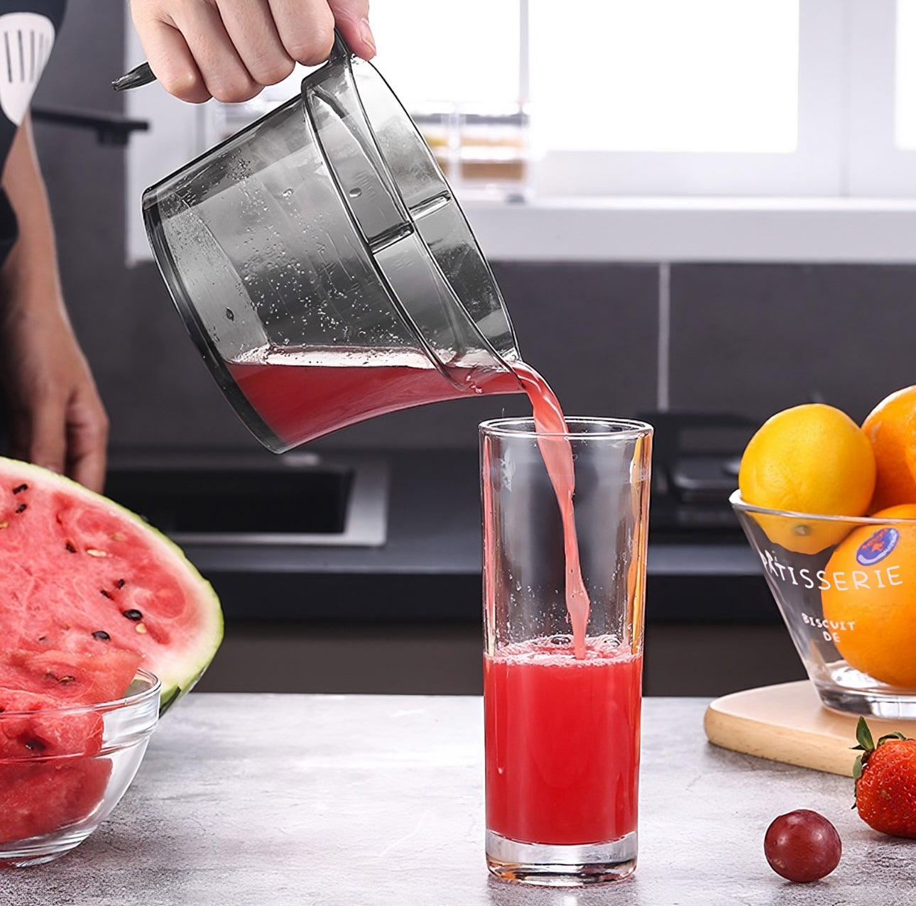 Hånd som heller rød fruktjuice fra sitruspresse i høyt glass på kjøkkenbenk med vannmelon og appelsiner.