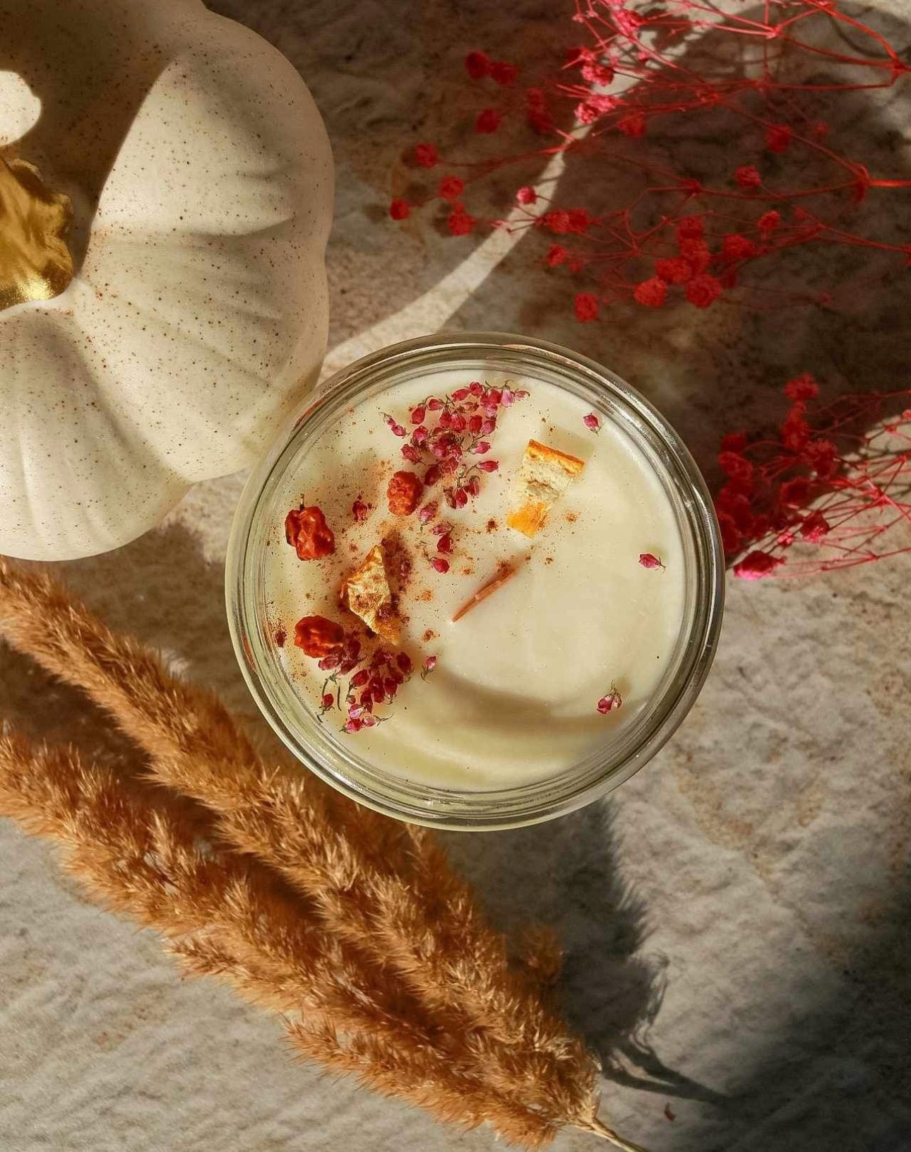 Soyaduftlys med tørket appelsin og blomster, omgitt av hvitt pyntegresskar og rosa tørkede blomster på steingulv