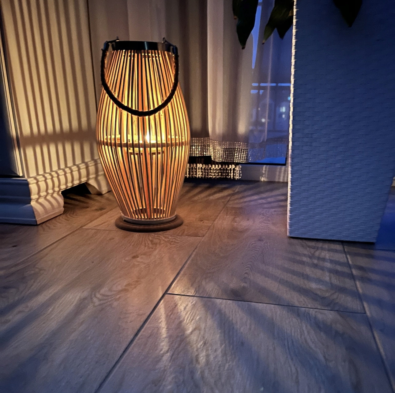 Bambuslyslykt med tent lys på gulv ved gardin, skaper varme skyggemønstre