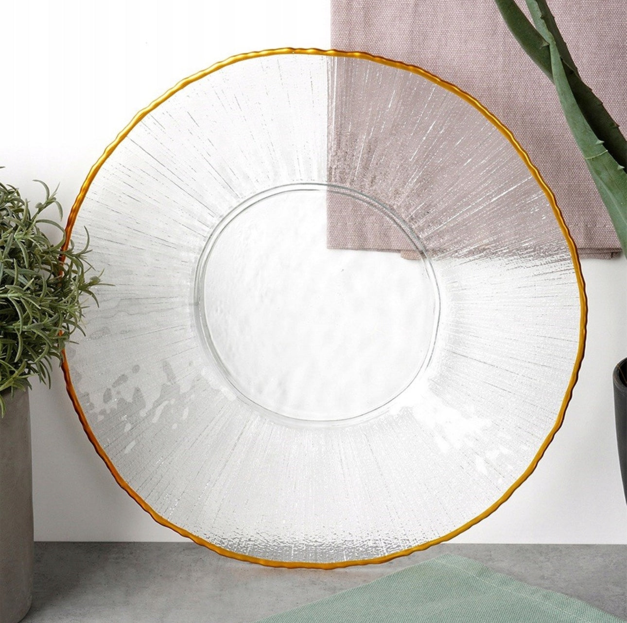 Tallerken i klart glass med gullkant plassert på spisebord med tekstiler og planter