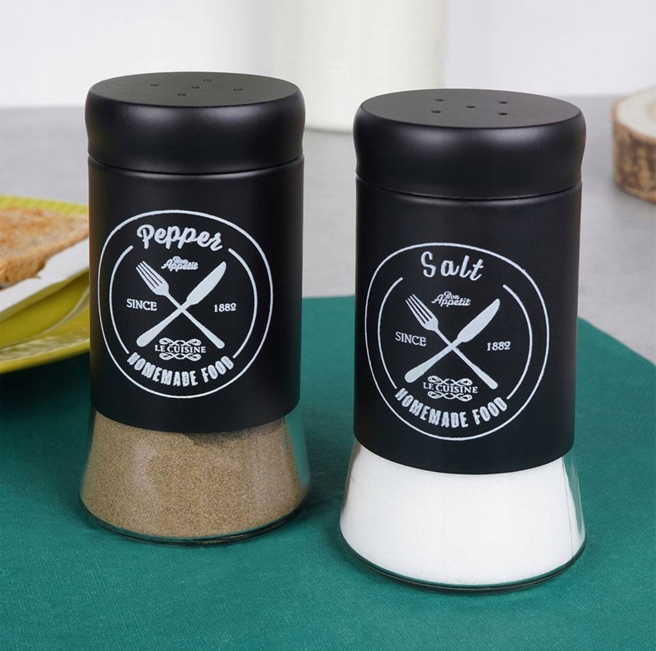 Salt- og pepperbøsser fylt med hvitt salt og brunt pepper, plassert på grønn duk ved siden av frokosttallerken.